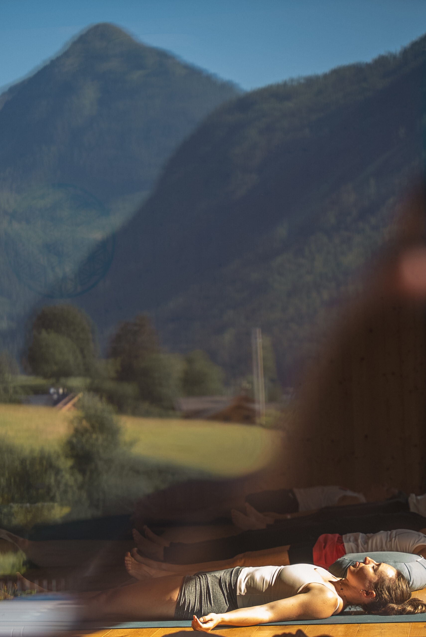 Gespiegelt im Glas ist eine Berglandschaft. Es wird in einen Yogaraum hineinfotografiert. PErsonen liegen in der Entenspannung am Boden.