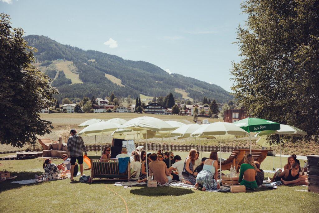 Viele Menschen bei einem Picknick unter Schirmen auf der Wiese vor Bergkulisse